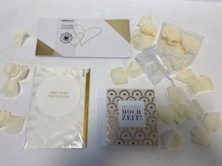 Weiß-gelbe Rosenblätter in weißem Organzasackerl, verschiedene Hochzeits-Billetts und Geschenkumschlag mit unterschiedlichen Motiven.