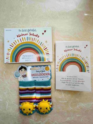 Meilensteine Affirmationskarten von Vera Lörks mit der Aufschrift Du bist geliebt, kleiner Schatz; bunt gestreifte Rasselsöckchen mit kleiner Sonne; Billett mit der Aufschrift Du bist geliebt, kleiner Schatz.